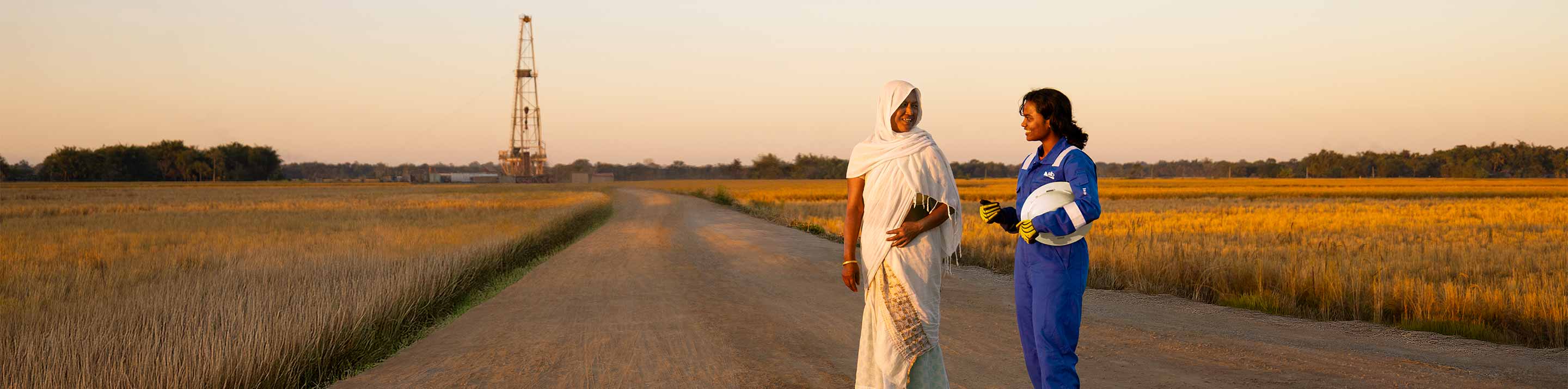 Two people on a road with an oil rig in the background. One is wearing blue SLB coveralls.