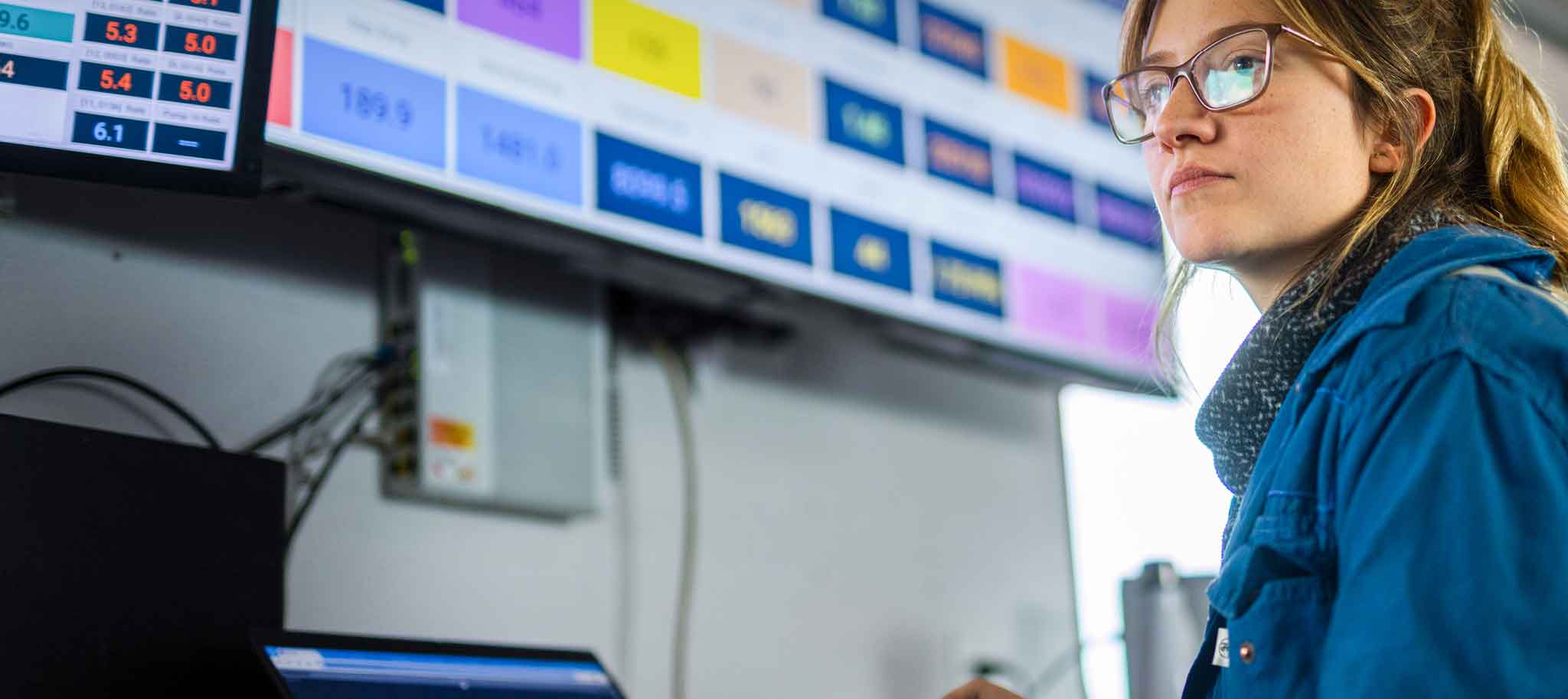 A woman wearing glasses and a blue jacket monitors multiple screens displaying data 