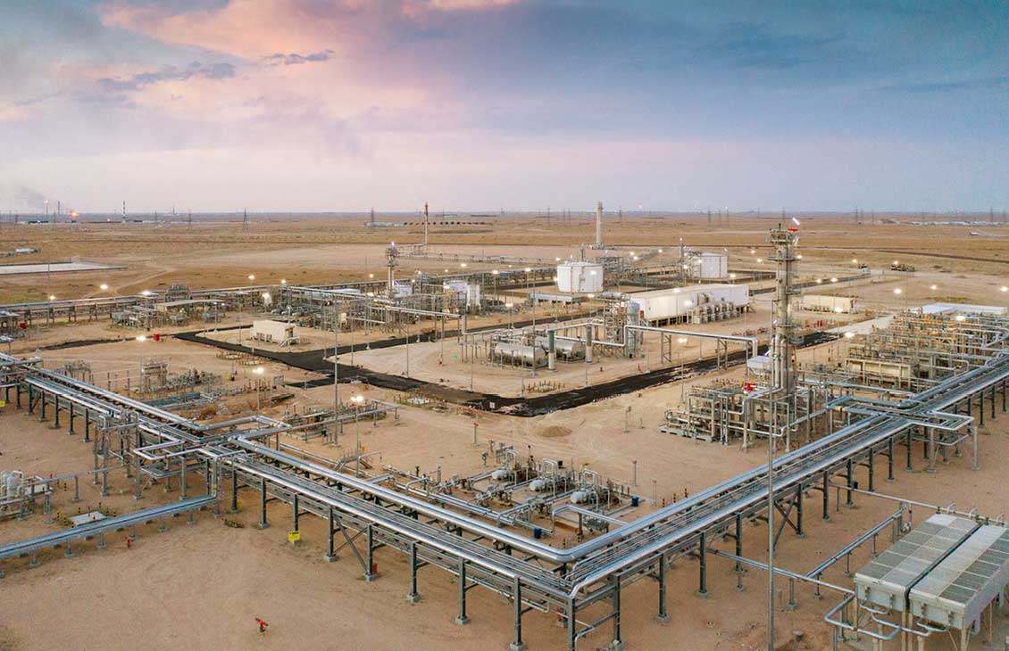 Aerial view of a large oil and gas processing facility in a desert landscape, with interconnected pipelines, storage tanks, and industrial structures under a colorful evening sky.